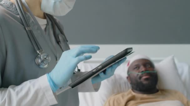 病院の部屋で負傷した黒人の健康状態をチェックしながら デジタルタブレットを使用して認識できない実践者の選択的な焦点 — ストック動画