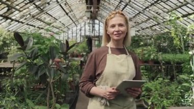 Serada elinde dijital tabletle kameraya gülümseyen beyaz önlüklü yaşlı bir kadının orta boy portresi.