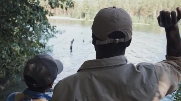 夏天的一天 一个难以辨认的非洲裔美国老人和他的孙子在湖边放松 用面包喂野鸭的背影 — 图库视频影像