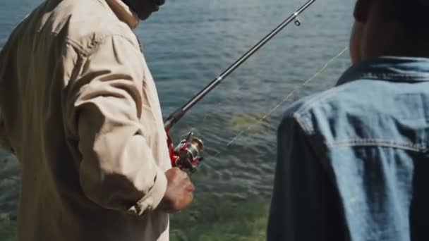 アフリカ系アメリカ人のシニアマンが孫に餌とフックで釣り棒をキャストする方法を教える選択的な焦点ショット — ストック動画