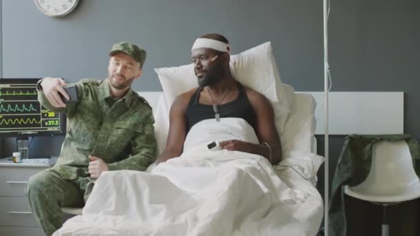 軍病院の部屋でスマートフォンで負傷した黒人の友人とセルフィーを取る陽気な白人兵士 — ストック動画
