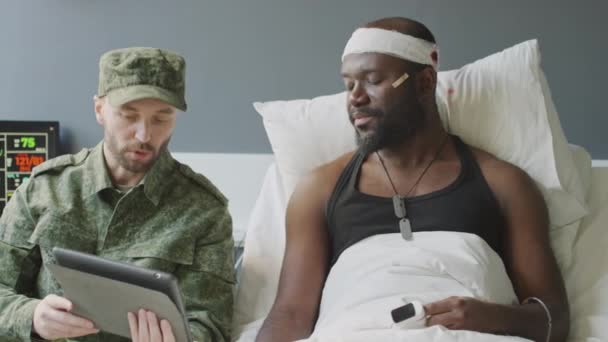 アフリカ系アメリカ人兵士の頭部負傷と彼の白人友人が軍病院で彼を訪問し デジタルタブレットで何かを見ています — ストック動画