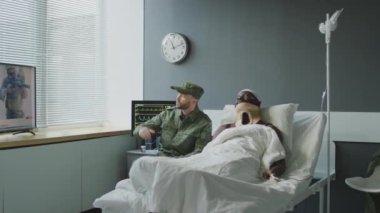 Kafasından ve boynundan yaralanmış Afrikalı Amerikalı bir adam ve hastane odasında televizyonda savaş haberleri izleyen Kafkasyalı bir asker.