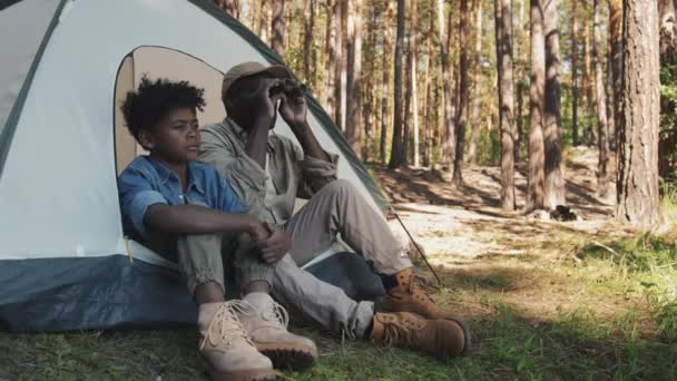 近代アフリカ系アメリカ人のシニアマンと孫がキャンプテントに座り 双眼鏡を使って遠くの何かを見つめている — ストック動画