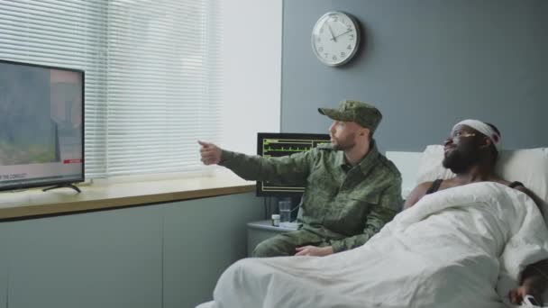 アフリカ系アメリカ人兵士の頭部負傷と彼のコーカサス人の友人が軍病院の部屋でテレビでニュースを見ている — ストック動画