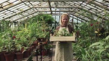 Kafkasyalı, önlük giymiş, elinde tahta kutuyla serasında dikilmiş kameraya gülümseyen yaşlı, neşeli bir kadın.
