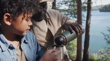 Afrika kökenli Amerikalı dedesinin göl kıyısında oturmuş torunu için vakumlu mataradan fincana çay dökerken orta boy fotoğrafı.