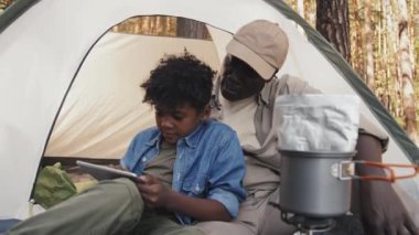 Orta boy Afro-Amerikan son sınıf öğrencisi ve torununun kamp çadırında oturup dijital masada bir şeyler izlediği bir fotoğraf.