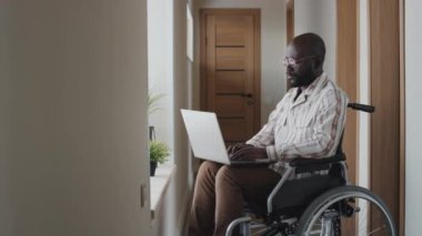 Orta boy, tekerlekli sandalyede, gözlüklü, koridorda oturmuş dizüstü bilgisayarda yazarken, muhtemelen çalışırken çekilmiş.