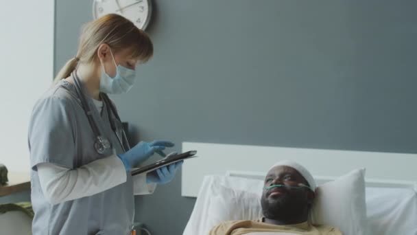女医生戴面具 用数码平板电脑填写医疗卡 与头部受伤的非裔美国男性病人交谈 — 图库视频影像