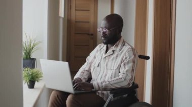 Orta boy, tekerlekli sandalyede oturan, daktiloyu bitiren, bilgisayarı kapatan ve kameraya bakan Afro-Amerikan bir adam.