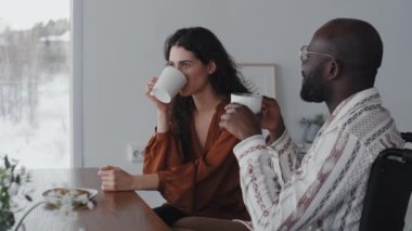Orta boy siyah adam ve beyaz kadın kahverengi bluzlu, açık oturma odasında kahve içen, birbirleriyle dostça konuşan, gülümseyen.