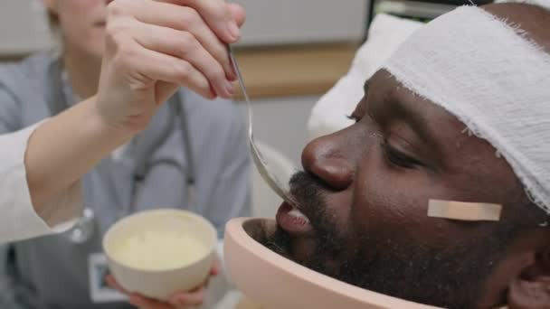 머리와 보조기에 붕대를 부상된 아프리카계 미국인 남자를 먹이는 간호사 숟가락의 — 비디오