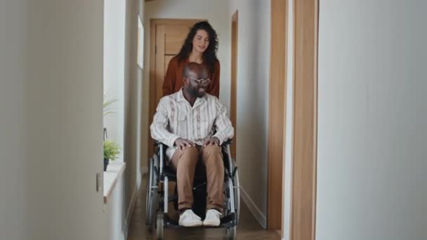 Black Man Riding Wheelchair Corridor Camera Smiling Assited Young Caucasian — Stock Video