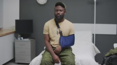 Orta boy Afro-Amerikan askerinin kol askısı taktığı hastane odasında kameraya bakarken.