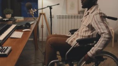 Tekerlekli sandalyedeki siyahi adamın kayıt sistemiyle masasına oturduğu, kulaklıklarını taktığı, daktilo etmeye başladığı, yan görüş aldığı orta boy bir fotoğraf.