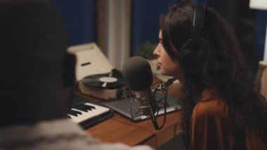 Kafkasyalı kadının kulaklıkla bir şey kaydettiği, mikrofona doğru konuştuğu, yanındaki siyahi adama baktığı görüntüler.