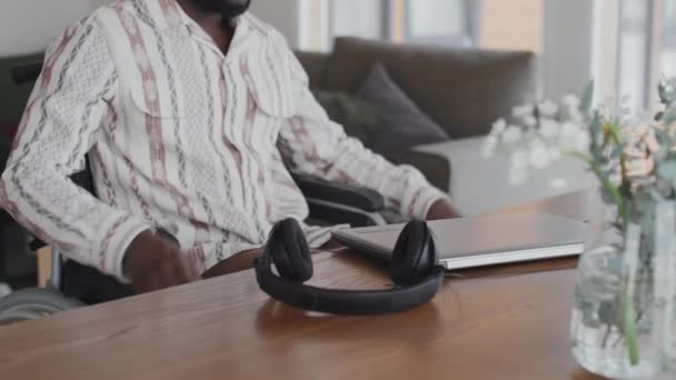 Чорний Чоловік Їде Інвалідному Кріслі Столу Відкриває Ноутбук Надягає Навушники — стокове відео