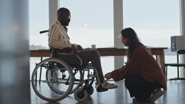 車椅子の黒人の男性のための靴ひもを結ぶ白人女性のミディアムフルショットは リビングルームからゆっくりと彼を運転 — ストック動画
