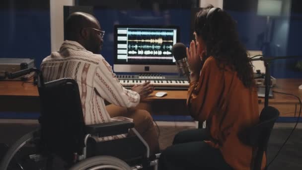 彼女と感情的に話す車椅子の黒人の男が彼女のヘッドフォンでオーディオを録音する中白人女性のミディアムフルショット — ストック動画