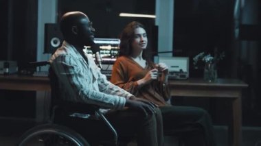 Orta boy, tekerlekli sandalyedeki Afrikalı Amerikalı adamın beyaz bir kadınla el ele tutuşup gece birlikte film izlerken gülümsediği bir fotoğraf.