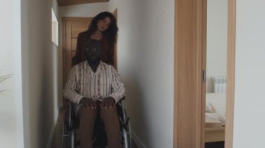 Kafkasyalı bir kadının siyah bir adamı tekerlekli sandalyeyle kameraya doğru sürdüğü orta boy bir fotoğraf. Adam ellerini dizlerinin üstünde tutup pencereden dışarı bakıyor.