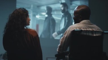 Orta boy beyaz kadın ve siyah adamın tekerlekli sandalyede büyük ekranda film izlerken, el ele tutuşurken ve birbirlerine gülümserken.