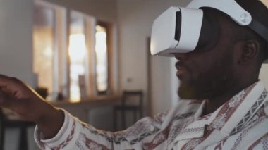 Beyaz VR kulaklık takmış, evde otururken sağ elini hareket ettiren siyah adamın yakın çekimi.