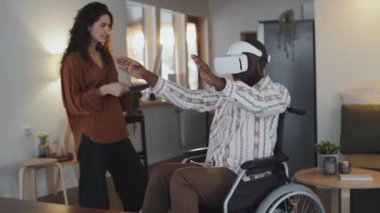 Orta boy tekerlekli sandalyede beyaz VR kulaklık takan siyah bir adam ellerini baş aşağı hareket ettirirken beyaz bir kadın yanında durup bilgisayarın tuşlarına basıyor ve gülümsüyor.