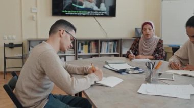 Orta Doğulu ve Siyahi göçmen öğrenciler İngilizce öğreniyorlar modern sınıfta masada oturmuş kompozisyon yazıyorlar.