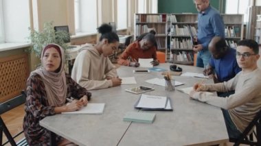 Başörtüsü takan Müslüman kadın ve çok ırklı grup arkadaşları masada oturmuş göçmenlere yönelik dil dersleri sırasında yazma görevi üzerinde çalışıyorlar