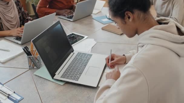 年轻的黑人男子和他的多种族同学坐在桌旁 一边看笔记本电脑上的视频 一边在抄袭本上记笔记 — 图库视频影像