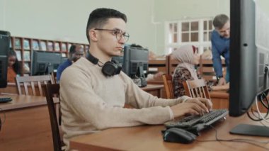 Gözlüklü ciddi bir Ortadoğu öğrencisi kütüphanede masaüstü bilgisayarında çalışıyor.