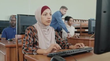 Genç Müslüman kadın tesettüre oturmuş ders sırasında masaüstü bilgisayarında bir şeyler yazıyor.