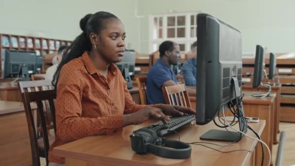 ヘッドフォンを置く大学図書館でデスクトップコンピュータを使用した現代アフリカ系アメリカ人女性学生のミディアムポートレート — ストック動画