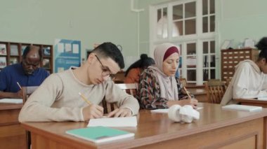 Etnik çeşitliliğe sahip genç ve olgun göçmen öğrencilerden oluşan bir grup sınıfta oturmuş ders sırasında notlar alıyorlar.