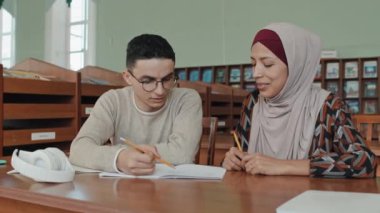 Orta Doğulu genç erkek ve kadın tesettürde oturmuş üniversite kütüphanesinde notlar okuyor ve görevlerini tartışıyorlar.