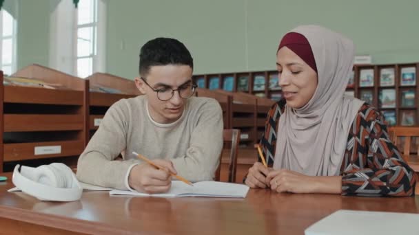 中东青年男女头戴头巾坐在大学图书馆的书桌前 阅读抄本上的笔记 讨论任务 — 图库视频影像