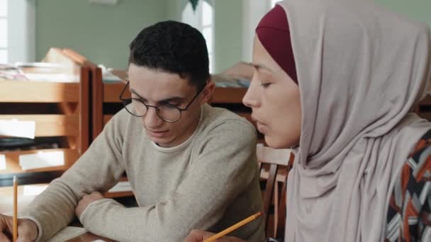 ヒジャブの若い中東人男性とムスリム女性の選択的な焦点ショットは 教室や図書館で一緒に文法演習をしているデスクに座っています — ストック動画