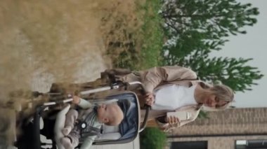 Dikey çekimde, beyaz kadın günlük giysiler içinde bebeğiyle dışarıda yürüyor.