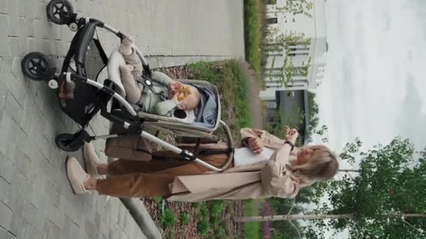 白种人年轻母亲穿着休闲装站在户外 带着孩子坐在婴儿车里 用智能手表记录语音信息的垂直全长镜头 — 图库视频影像