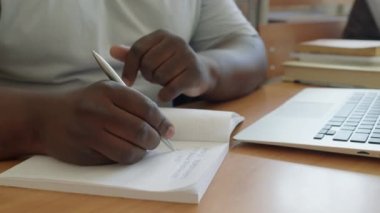 Tanımlanamayan siyahi erkek üniversite öğrencisi masada oturmuş defterine not alıyor ve dizüstü bilgisayar kullanıyor.