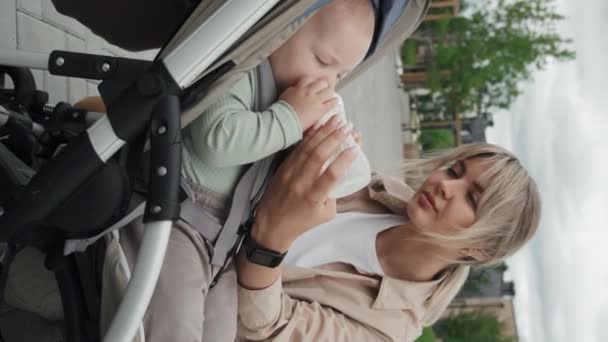 屋外で時間を過ごしながらベビーカーで彼女の赤ちゃんの息子にボトルで公式を与える若い白人母親の垂直ショット — ストック動画