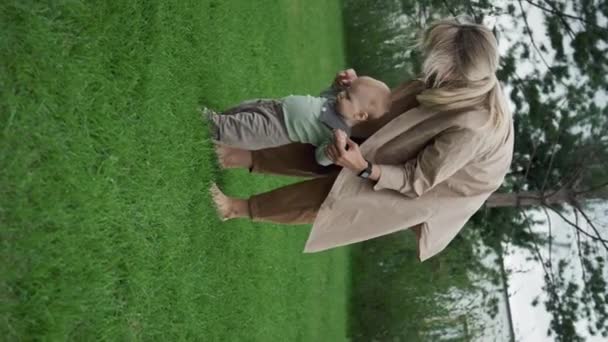 赤ちゃんに歩くように教える現代の若い母親の垂直ショット 芝生の草の上に裸足を歩く — ストック動画