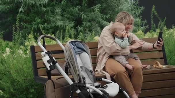 幸せな若い白人母親は 彼女のラップレコーディングビデオ上の赤ちゃんと公園でベンチに座っているか スマートフォンで写真を撮る — ストック動画