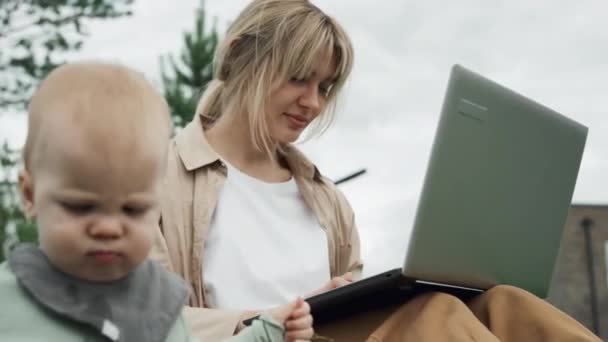 年轻母亲坐在草坪上用笔记本电脑看着她的小儿子的低角度选择性聚焦镜头 — 图库视频影像
