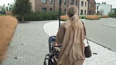 Sarı saçlı, günlük kıyafetler giyen, bebek arabasıyla dışarıda yürüyen genç annenin fotoğrafını takip et.