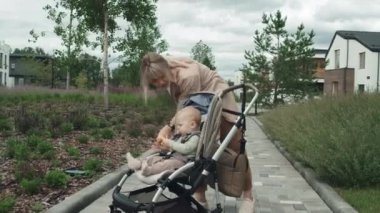 Kafkasyalı genç bir kadın günlük kıyafetlerini giymiş dışarıda bebeğiyle birlikte duruyor bebek arabasında oturuyor ve akıllı saat kullanarak sesli mesaj kaydediyor.