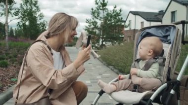 Mutlu genç bir kadının bebek arabasında akıllı telefonuyla neşeli oğlunun fotoğrafını çektiği orta boy bir fotoğraf.