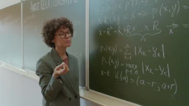 Matematik öğretmeninin orta boy portresi tahtaya oturmuş denklemi nasıl çözeceğini anlatıyor.